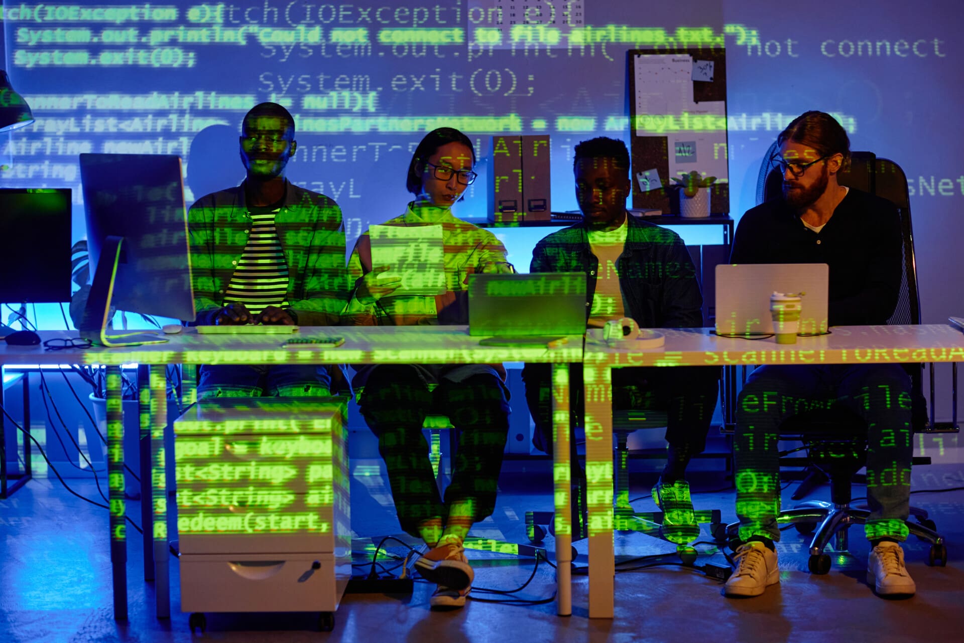 Row of four young diversity programmers sitting by desk in front of laptops